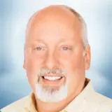A middle-aged man with light skin, a bald head, and a white goatee smiles at the camera. He wears a light-colored collared shirt against a soft blue gradient background, conveying warmth and approachability. Sourcing, Procurement, 3rd Party Risk