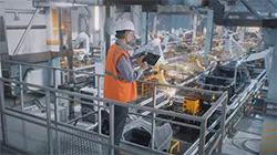 A worker in a white hard hat and orange safety vest inspects a tablet on an elevated platform in a modern, automated factory with robotic arms and advanced machinery below. Sourcing, Procurement, 3rd Party Risk