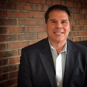 A man in a dark blazer and light shirt smiles while seated in front of a red brick wall, exuding confidence. Sourcing, Procurement, 3rd Party Risk