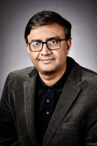 A man with short dark hair and glasses, wearing a black collared shirt and a dark textured blazer, smiles slightly while posing against a neutral gray studio background. Sourcing, Procurement, 3rd Party Risk