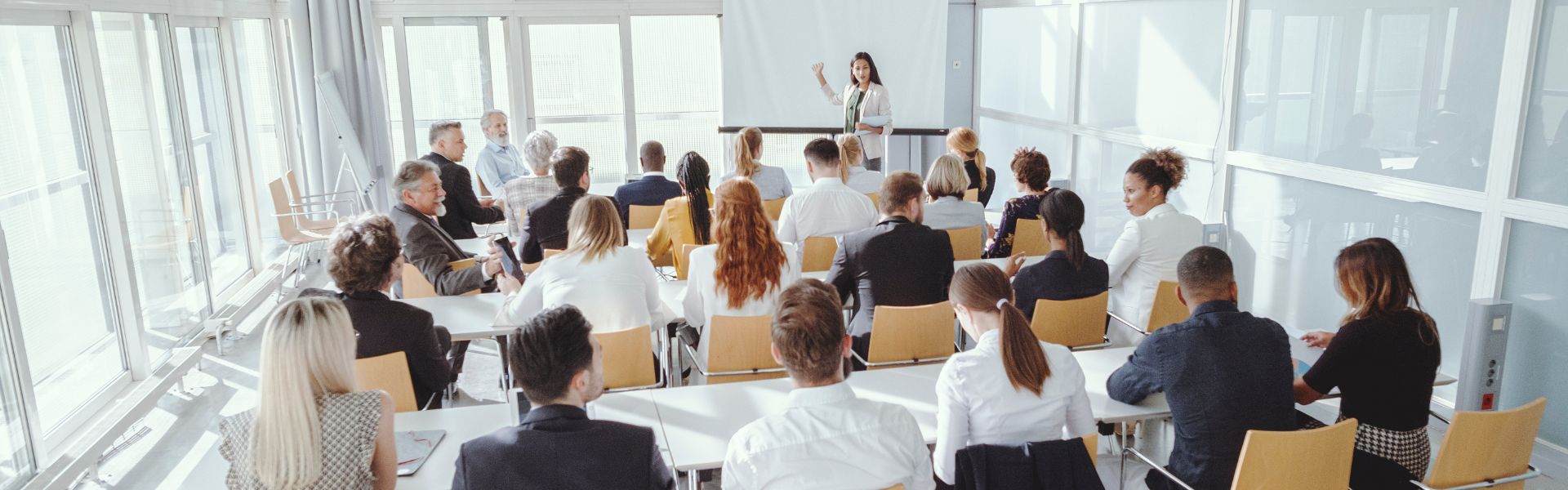 A group of people sit in rows facing a speaker at the front of a bright, modern conference room. The speaker stands near a whiteboard, engaging the attentive audience in a dynamic presentation. Sourcing, Procurement, 3rd Party Risk