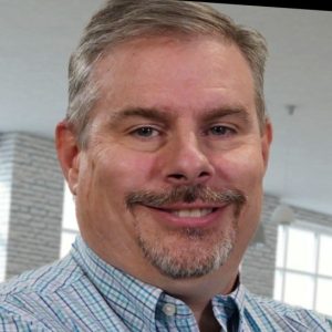 A middle-aged man with short gray hair and a goatee smiles at the camera, wearing a plaid shirt. Natural light streams through bright windows, highlighting the brick wall in the softly blurred background. Sourcing, Procurement, 3rd Party Risk