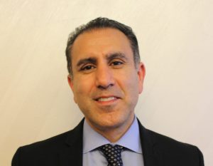 A man wearing a dark suit, light blue dress shirt, and patterned tie smiles confidently at the camera against a neutral background. Sourcing, Procurement, 3rd Party Risk