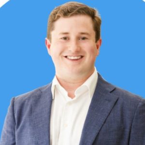 A smiling man with short brown hair is wearing a white dress shirt and a stylish blue blazer, standing in front of a vibrant solid blue background. Sourcing, Procurement, 3rd Party Risk