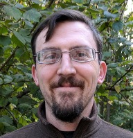 A man with glasses, short brown hair, and a beard is smiling outdoors. He is wearing a brown zip-up sweater, and leafy green trees are visible in the background under natural light. Sourcing, Procurement, 3rd Party Risk