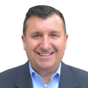 A man with short dark hair is smiling in a dark suit jacket over a light blue shirt, standing against a plain white background, exuding professionalism. Sourcing, Procurement, 3rd Party Risk