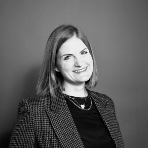 A woman with straight, shoulder-length hair smiles at the camera. She is wearing a checked blazer over a dark top and layered necklaces. The plain, neutral background enhances the black-and-white photo's timeless style. Sourcing, Procurement, 3rd Party Risk