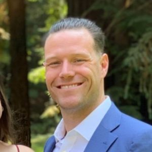 A man with short, slicked-back hair wearing a blue suit and white shirt is smiling outdoors in a sunlit, wooded landscape. Sourcing, Procurement, 3rd Party Risk