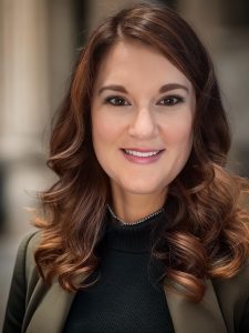 A woman with long, wavy brown hair is smiling. She wears a dark top with a blazer and a beaded necklace. The softly blurred background suggests an urban setting, highlighting her confident presence. Sourcing, Procurement, 3rd Party Risk