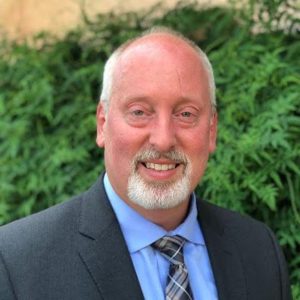 A smiling bald man with a white beard, wearing a dark suit, blue shirt, and plaid tie, stands outdoors in front of lush green bushes. Sourcing, Procurement, 3rd Party Risk
