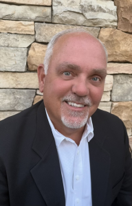 A smiling middle-aged man with short gray hair and a goatee, dressed in a white shirt and black blazer, stands in front of a wall made of stacked stone bricks, exuding confidence. Sourcing, Procurement, 3rd Party Risk