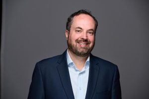 A man with short dark hair and a beard, wearing a navy blue suit jacket and light blue shirt, confidently smiles at the camera against a plain gray background. Sourcing, Procurement, 3rd Party Risk
