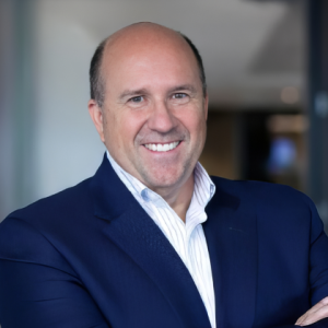 A middle-aged man in a dark blue suit and white striped shirt smiles confidently with arms crossed in a modern office setting. Sourcing, Procurement, 3rd Party Risk