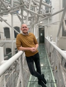 A man in a mustard sweater and black jeans leans against a railing on a glass walkway with white metal pipes overhead in a modern, industrial loft. Sourcing, Procurement, 3rd Party Risk