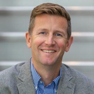 A man with short light brown hair smiles at the camera, wearing a gray blazer over a blue and white checkered shirt. The blurred background features horizontal steps or lines. Sourcing, Procurement, 3rd Party Risk