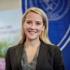A woman with long blonde hair, wearing a dark blazer and gray top, smiles at the camera. She stands indoors in front of a blurred blue and white background featuring a partial emblem. Sourcing, Procurement, 3rd Party Risk