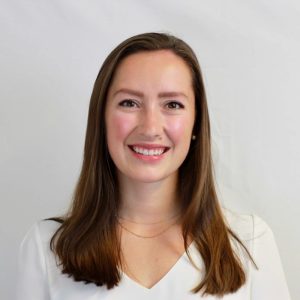 A woman with straight brown hair and fair skin smiles at the camera. She is wearing a white top, a delicate gold necklace, and stands in front of a plain light background. Sourcing, Procurement, 3rd Party Risk