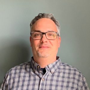 A man with short gray hair and glasses, wearing a checked button-up shirt, stands in front of a plain gray background, smiling slightly at the camera. Sourcing, Procurement, 3rd Party Risk