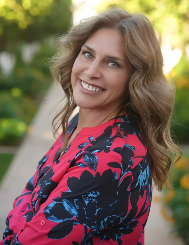 A woman with wavy light brown hair smiles at the camera in a vibrant pink and blue floral top, standing outdoors amidst lush greenery and a sunlit garden path. Sourcing, Procurement, 3rd Party Risk