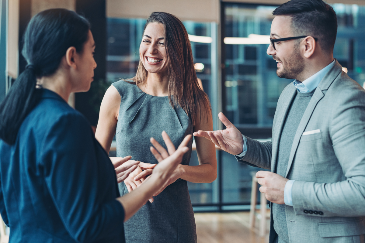 Three professionally dressed people stand together indoors, engaged in a lively conversation. One woman is smiling broadly while the other woman and a man gesture enthusiastically, showcasing strong teamwork. Sourcing, Procurement, 3rd Party Risk