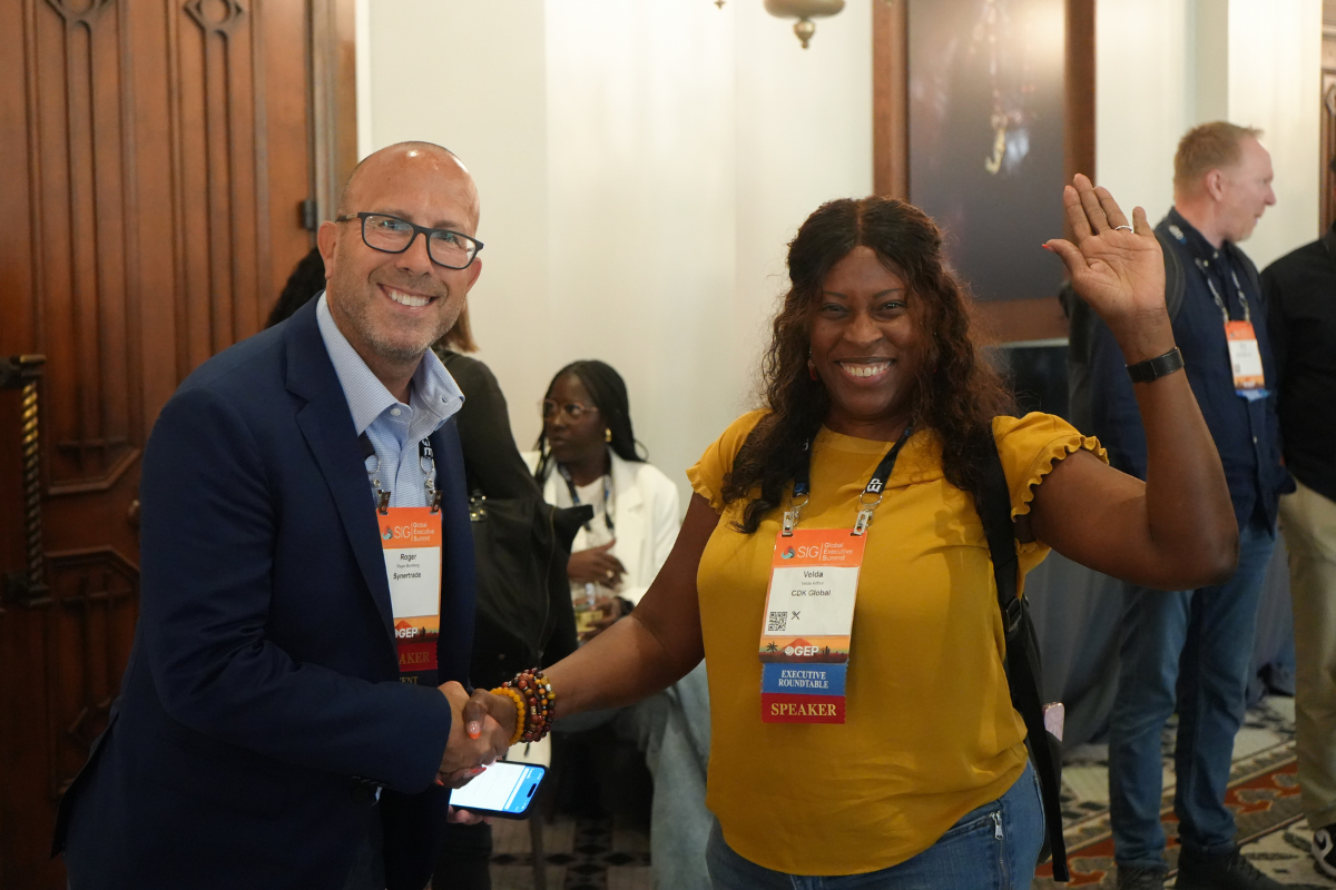 Two people at a conference smile and shake hands, with the woman on the right waving. Both wear name tags as other attendees are seated in the background. The atmosphere is friendly and professional. Sourcing, Procurement, 3rd Party Risk