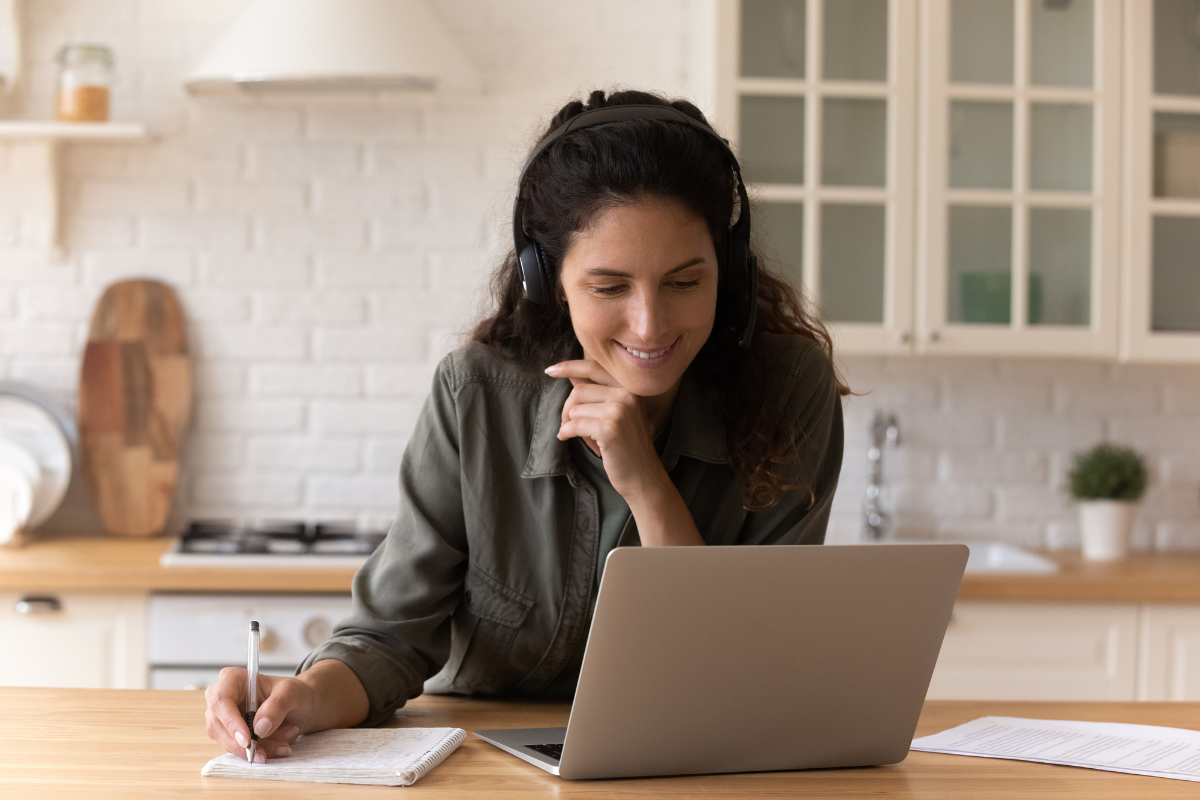 A woman wearing headphones sits at a kitchen counter, smiling at her laptop. She writes in a notebook, suggesting she’s studying or working remotely. The scene features a light, modern kitchen background. Sourcing, Procurement, 3rd Party Risk
