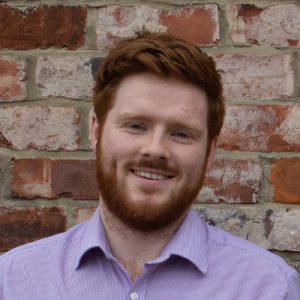 A smiling man with red hair and a beard, wearing a light purple shirt, stands in front of a rustic brick wall. Sourcing, Procurement, 3rd Party Risk