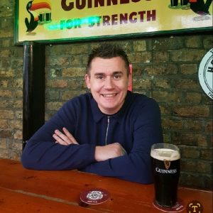 A man with short brown hair, wearing a navy blue sweater, sits at a wooden table with a pint of Guinness in front of him. He is smiling, and there’s a brick wall and an illuminated Guinness sign in the background. Sourcing, Procurement, 3rd Party Risk