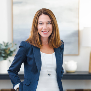A woman with straight, shoulder-length brown hair in a blue blazer and white top stands indoors, smiling at the camera. A blurred painting and a green plant are visible in the background. Sourcing, Procurement, 3rd Party Risk