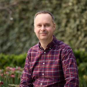 A man with short hair wearing a red and blue plaid shirt sits outdoors, smiling at the camera. In the background, lush green bushes and blurred plants create a serene atmosphere. Sourcing, Procurement, 3rd Party Risk