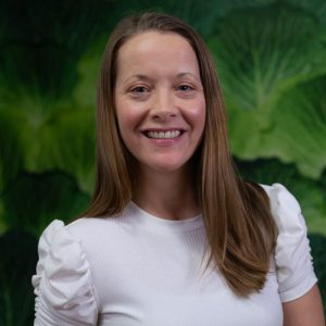 A woman with straight, long light brown hair wearing a white short-sleeved top stands smiling in front of lush greenery. Sourcing, Procurement, 3rd Party Risk
