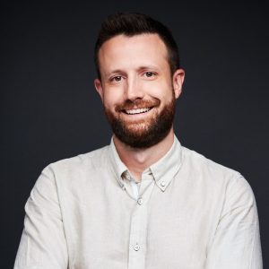 A man with short brown hair and a beard, wearing a light-colored button-up shirt, confidently smiles with arms crossed in front of a dark background. Sourcing, Procurement, 3rd Party Risk