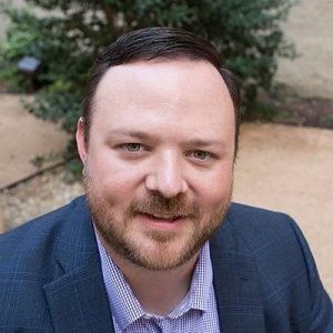 A man with short dark hair and a beard, wearing a blue checked blazer and a light purple shirt, smiles at the camera outdoors amid lush greenery and a beige wall in the background. Sourcing, Procurement, 3rd Party Risk