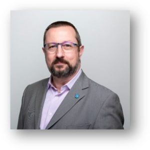 A middle-aged man with short hair, a beard, and glasses, wearing a gray suit jacket over a light purple shirt, poses confidently in front of a plain light background. Sourcing, Procurement, 3rd Party Risk