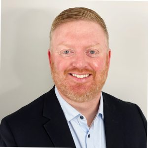 A man with short, light brown hair and a reddish beard, wearing a black blazer and light blue collared shirt, smiles warmly at the camera against a plain background. Sourcing, Procurement, 3rd Party Risk