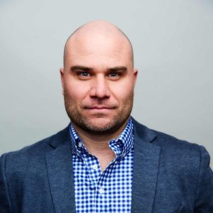 A bald man with a trimmed beard, wearing a blue blazer and blue-and-white checkered shirt, looks confidently at the camera against a plain light gray background. Sourcing, Procurement, 3rd Party Risk