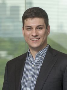 A young man in a checked shirt and dark blazer smiles at the camera, standing indoors with a blurred window and greenery in the background, exuding confidence. Sourcing, Procurement, 3rd Party Risk