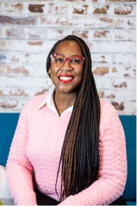 A smiling woman with long braided hair and red glasses wears a pink sweater over a white collared shirt, sitting in front of a textured white brick wall. Sourcing, Procurement, 3rd Party Risk