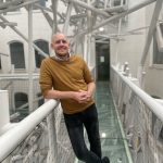 A person in a mustard sweater and black jeans leans on a glass walkway surrounded by modern white metal structures indoors, exuding confidence and smiling at the camera. Sourcing, Procurement, 3rd Party Risk