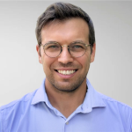 A man with short brown hair, glasses, and a light blue collared shirt is smiling at the camera against a plain light background. Sourcing, Procurement, 3rd Party Risk
