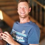 A smiling man with short light brown hair sits indoors, wearing a blue Volkswagen t-shirt and holding a smartphone. The blurred background reveals a staircase and wooden railing, highlighting his relaxed mood. Sourcing, Procurement, 3rd Party Risk
