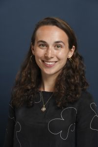 A woman with long curly brown hair, wearing a black top with white patterns, gold hoop earrings, and a gold necklace, smiles confidently in front of a sleek dark blue background. Sourcing, Procurement, 3rd Party Risk