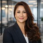 A woman with long, wavy brown hair, wearing a dark blazer and white top, smiles confidently while standing in a modern glass-walled office filled with bright lighting. Sourcing, Procurement, 3rd Party Risk