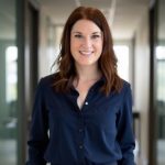 A woman with shoulder-length brown hair, wearing a navy blue button-up shirt, smiles while standing in a bright office hallway with glass walls. Sourcing, Procurement, 3rd Party Risk