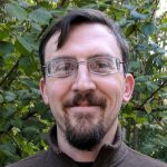 A man with short brown hair, glasses, and a beard is smiling at the camera. He is wearing a brown zip-up jacket, and green leaves and branches fill the background. Sourcing, Procurement, 3rd Party Risk