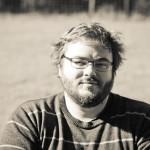 Black and white photo of a bearded person with glasses, wearing a striped sweater, sitting outdoors with a blurred natural background. Sourcing, Procurement, 3rd Party Risk