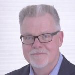 A middle-aged man with short gray hair, glasses, and a trimmed white beard and mustache, wearing a dark suit jacket and light blue shirt, smiles slightly in front of a bright, minimalist background. Sourcing, Procurement, 3rd Party Risk