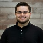 A man with short dark hair, a beard, and glasses smiles at the camera. He is wearing a black polo shirt and standing in front of a light brown and white brick wall. Sourcing, Procurement, 3rd Party Risk