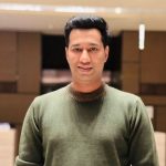 A man with short black hair, wearing a green sweater, stands indoors in a softly-lit room with a modern, brown-toned background. He is smiling and facing the camera. Sourcing, Procurement, 3rd Party Risk