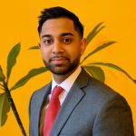 A man in a gray suit and red tie stands before a bright yellow wall with a leafy green plant beside him, smiling slightly at the camera, exuding confidence and professionalism. Sourcing, Procurement, 3rd Party Risk